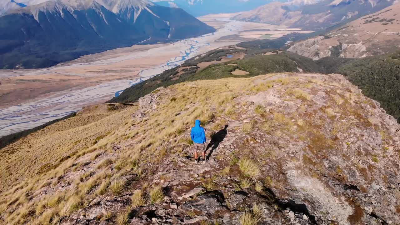 Man In Blue Hoodie Jacket Standing On The Mountain Ridge In Arthur's Pass National Park, Canterbury, New Zealand With Overview Of The Scenic Bealey Spur Track - orbiting drone shot