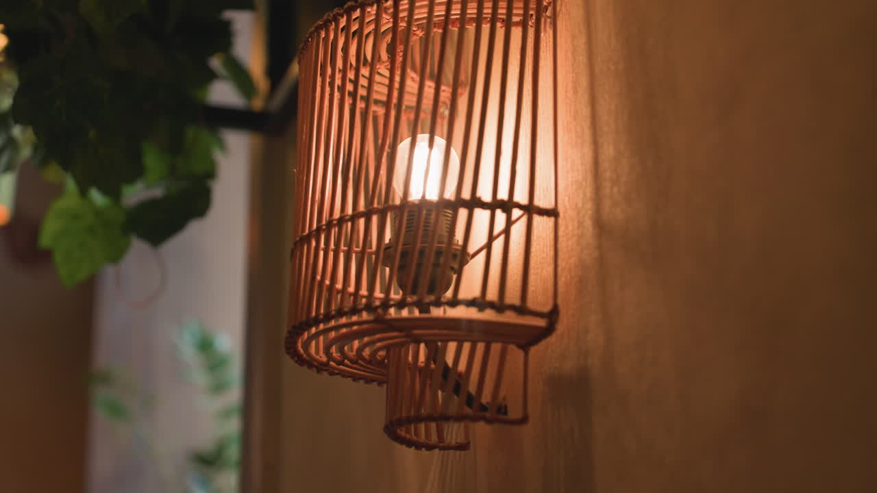 close up decorative wall lamp enclosed in wooden slats casting soft warm glow and shadow on wall with surrounding green leaves enhancing cozy and natural ambiance in indoor space