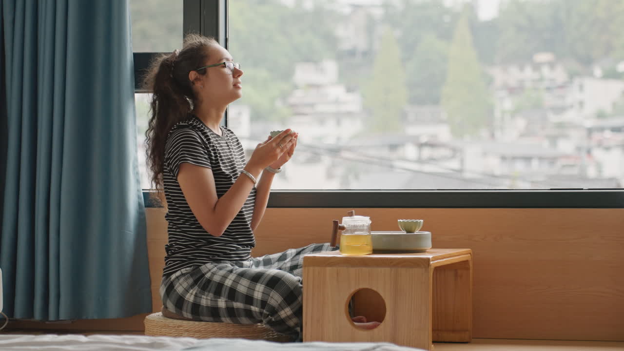 Donna in camera d'hotel che ringrazia, mani giunte sopra una tazza fumante su un vassoio di legno, skyline della città visibile attraverso il vetro, tende morbide, energia mattutina calma, viaggiatore consapevole che pratica la gratitudine.