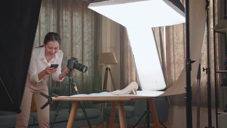 asian female photographer comparing photos on a smartphone to camera while taking photos of women's shoes in home studio