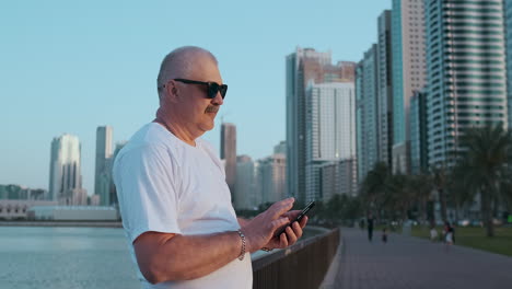 Low-angle-view-of-attractive-business-man-over-blurred-street-background-holding-smart-phone-in-hands-using-4G-internet-chatting-with-friends-searching-contact.-Electronic-wireless-device-concept.