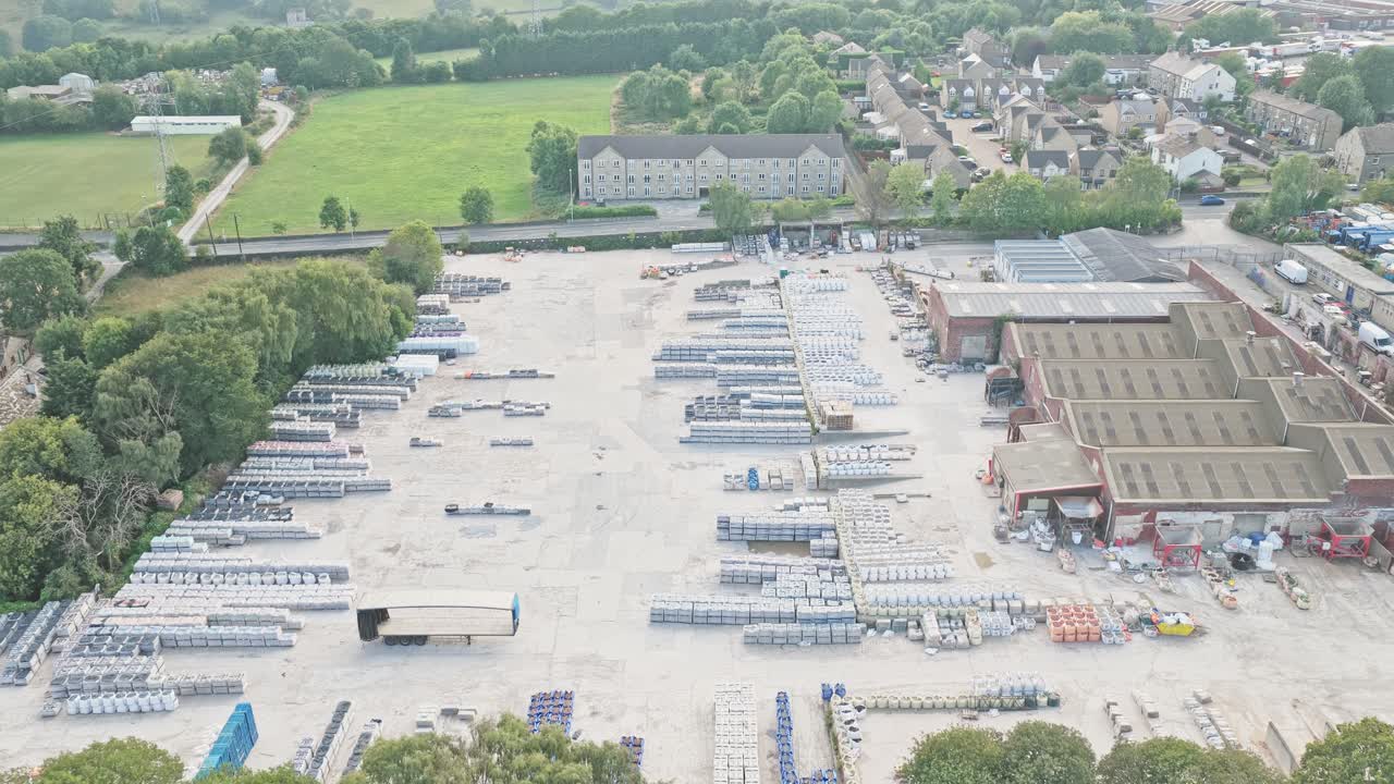 Drone pushes in on Sunderland Way industrial park, Brighouse, Calderdale, West Yorkshire, England, revealing stacked building materials, warehouses, and boundary trees