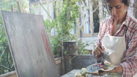 Biracial-female-artist-in-apron-painting-on-canvas-in-sunny-garden,-slow-motion