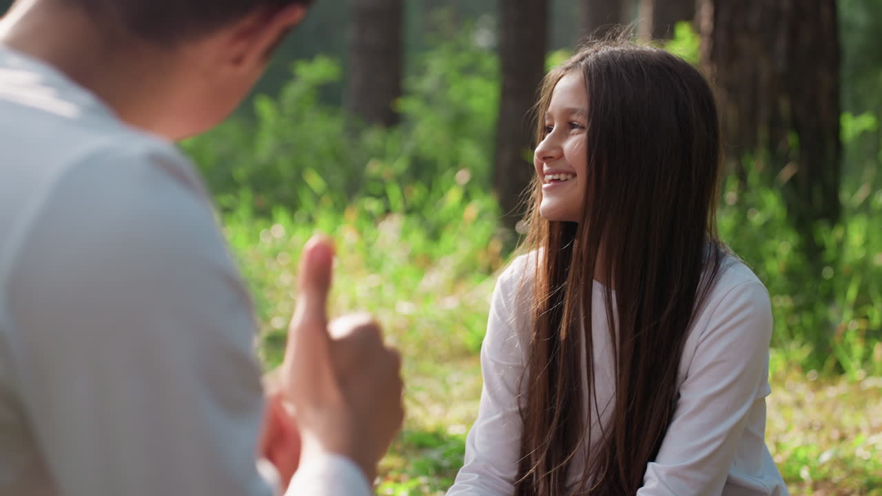Over shoulder view of big brother making gestures while talking to sister during picnic, young girl smiling warmly while listening in sunlight surrounded by forest greenery, peaceful family bonding