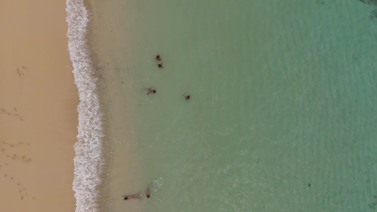 Ripresa a volo d'uccello con drone dall'alto su una spiaggia tropicale nel sud di Lombok, che mostra bambini che giocano in acque turchesi poco profonde, onde delicate, costa sabbiosa e atmosfera costiera tranquilla