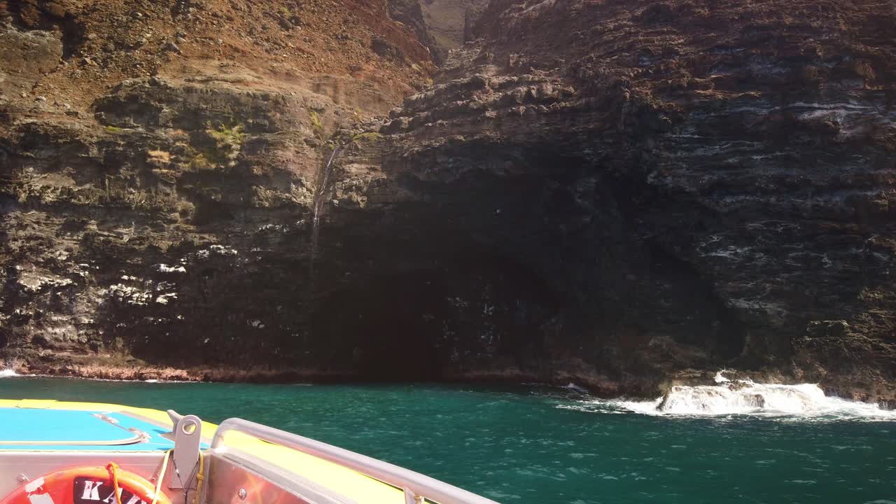 Gimbal wide POV shot from a moving boat approaching a sea cave with a waterfall cascading over the entrance along the southern Na Pali Coast in Kaua'i, Hawai'i