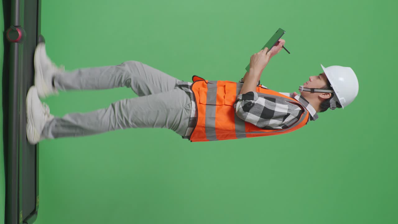 안전 헬을 입은 아시아 남성 엔지니어가 녹색 스크린 배경 스튜디오에서 는 동안 태블릿에 메모를 취합니다.