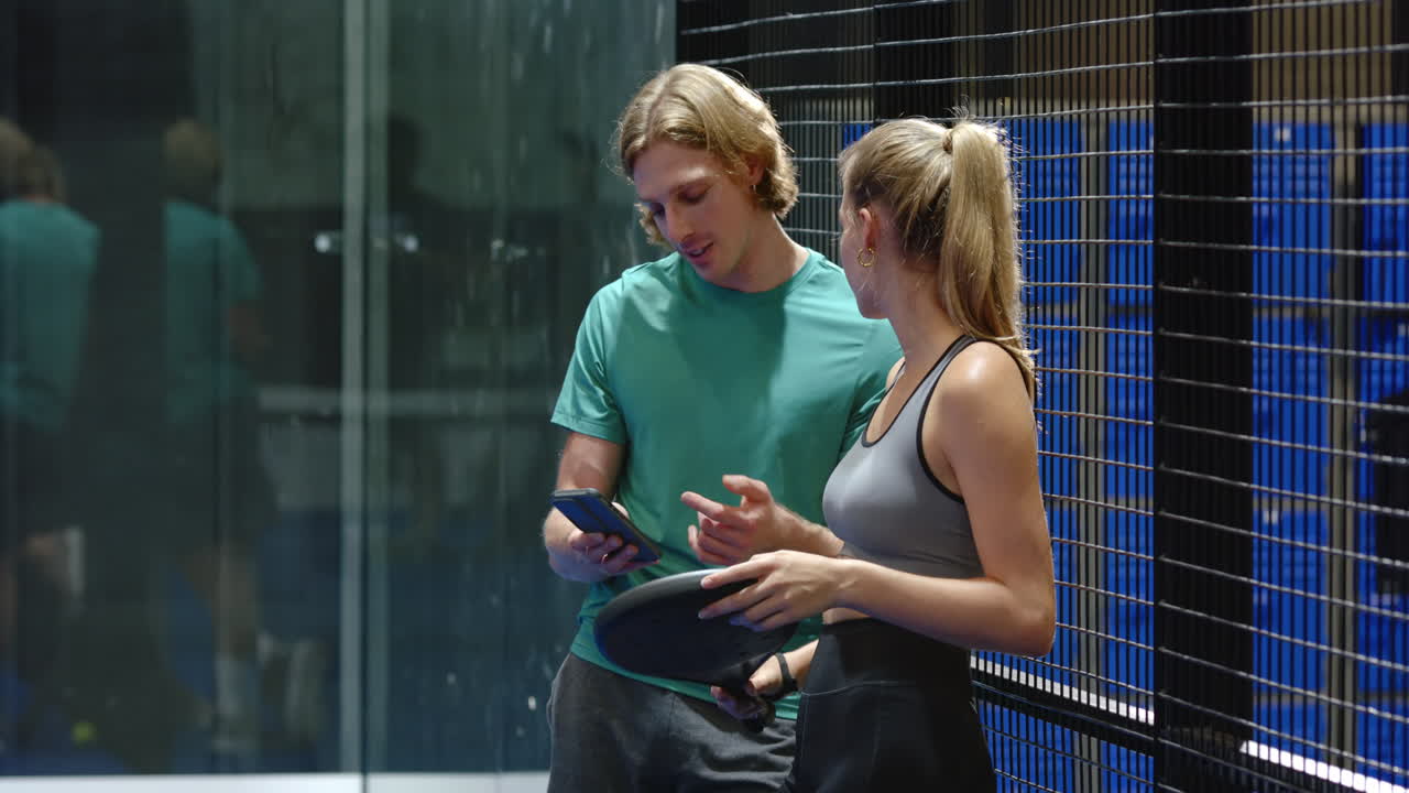 Man and woman on break from padel tennis, sharing smartphone, smiling, on indoor court, copy space