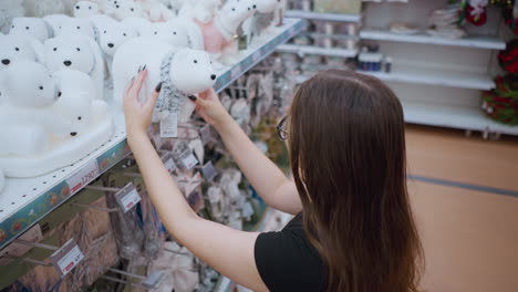 vista lateral de una mujer joven de cabello largo y marrón con camisa negra y gafas admirando un juguete de oso polar de peluche blanco con bufanda en la estantería de la tienda rodeada de decoraciones festivas y iluminación cálida