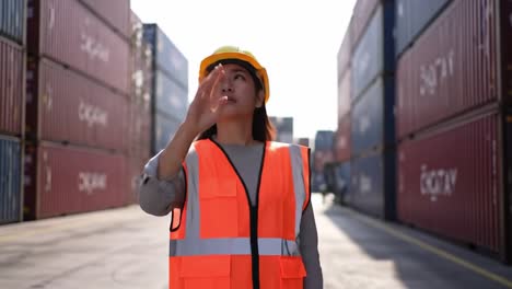 a female worker in a high-visibility vest and hard hat directing operations amidst stacks of shipping containers in a freight yard environment