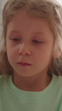 calm blonde long haired child in green sweatshirt taps finger playing at table against window closeup slow motion. happy childhood and development games