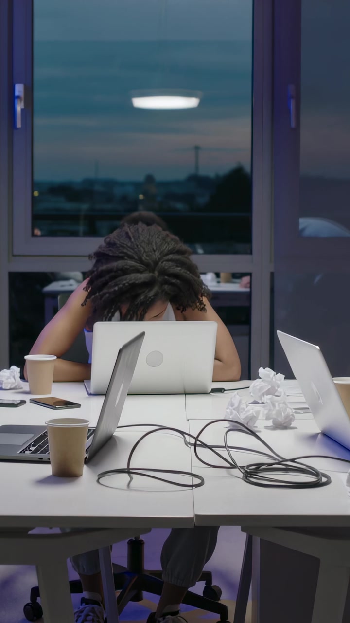 Vertical video: Leaning worker holding head on laptop at desk at dusk, wearing casual top