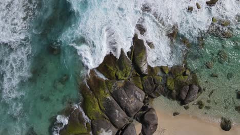 aerial top down shot of crashing waves against rocky and mossy stones at shore of ocean with sandy beach
