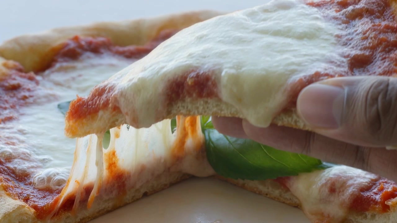 Mano destra che afferra la crosta e solleva una fetta di pizza dal piatto, rivelando il basilico e la mozzarella filante.