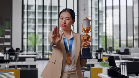 asian business woman in a suit with a gold medal and trophy disapproving with no hand sign and smiling to camera in the office