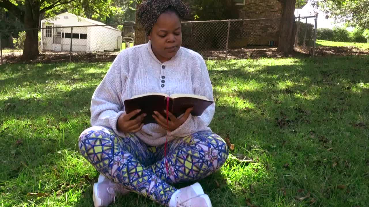 zoom lento de una mujer negra sentada con las piernas cruzadas en el patio de una casa suburbana leyendo su biblia y pasando tiempo con dios
