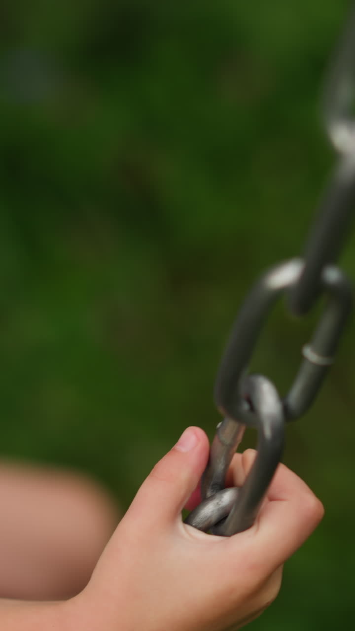 Preschooler fair haired girl in shorts sits on wooden swings with chains on playground on gloomy spring day upper close view slow motion