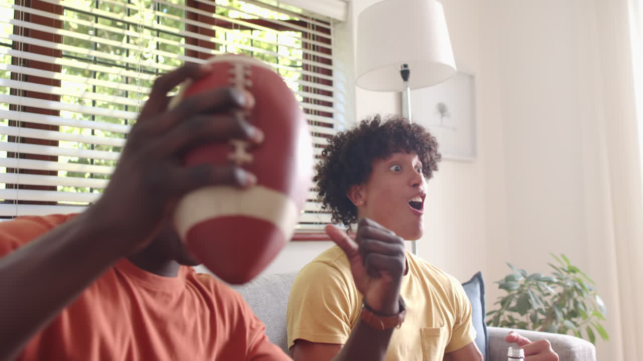 Cheering enthusiastically, two multiracial male friends holding football watching game, at home
