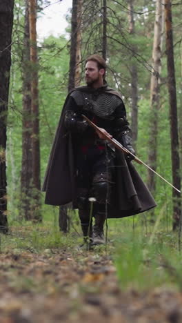 brave military man with cape and bow patrols forest slow motion. middle age bearded soldier with armor walks across woodland. historical camp