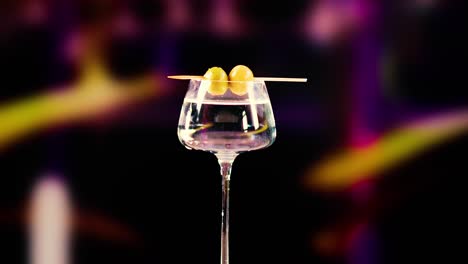 a hand places olives into a martini glass against a blurred, colorful background. the lighting highlights the glass and olives