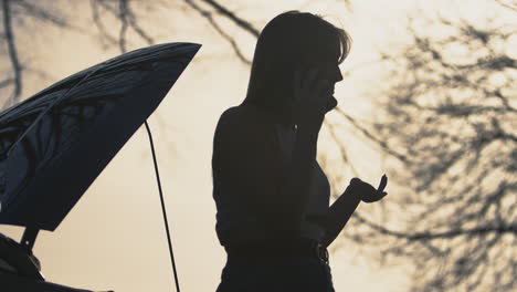 Silhouette-Einer-Frustrierten-Frau-Mit-Kaputtem-Auto-Auf-Einer-Landstraße,-Die-Per-Mobiltelefon-Um-Hilfe-Ruft