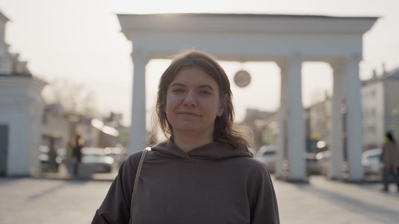 Donna tiene e manipola una cinghia di pelle marrone in un contesto urbano, indossando abiti casual con luce naturale soffusa. Lo sfondo presenta elementi architettonici come colonne sotto la luce del giorno.