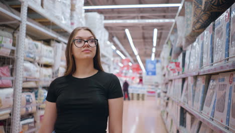 woman in black top walking near supermarket shelf focused on textile products display, with blurred background showing another shopper pushing cart in distance, bright retail environment