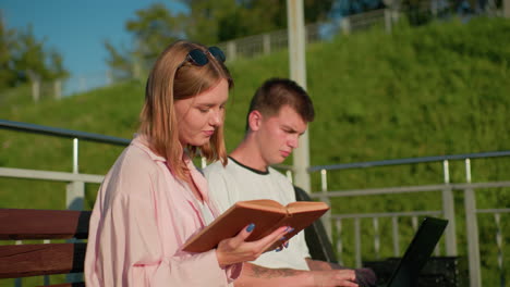 lady in pink reading from her book while her friend next to her types on a laptop and nods in agreement, background features green hill, pole, and outdoor bench in warm natural light