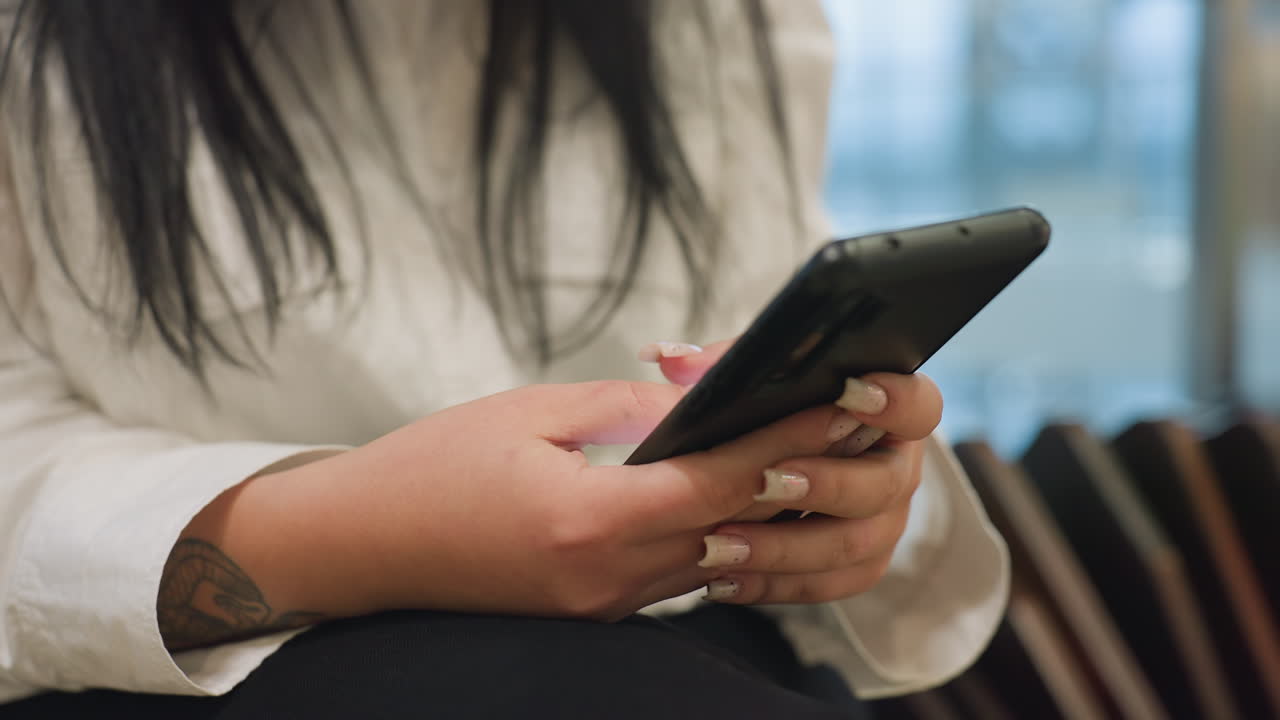 Close up side view of young woman in white shirt holding smartphone with both hands, seated indoors, fingers gently tapping screen, background softly blurred