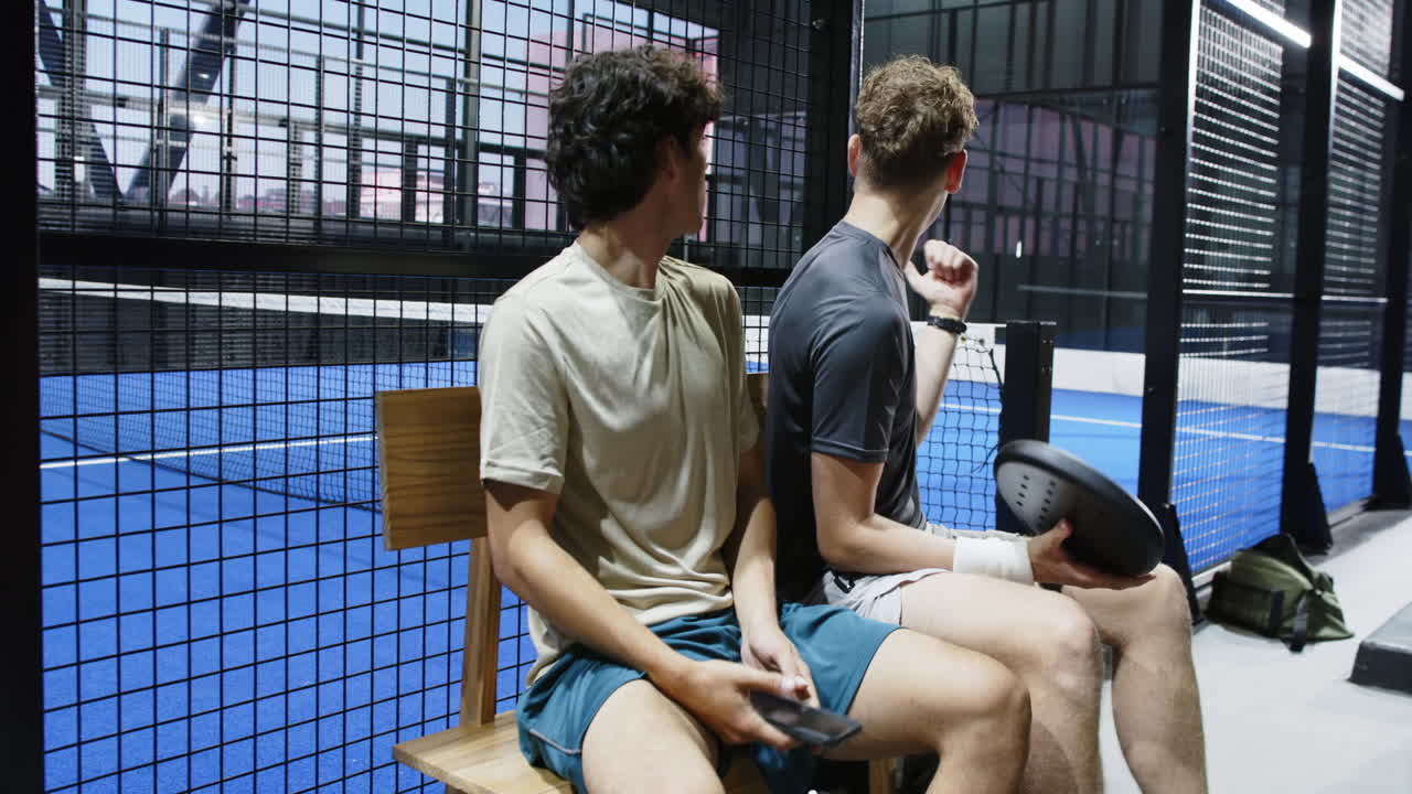 Young men sitting on bench, holding padel racket, enjoying conversation at court