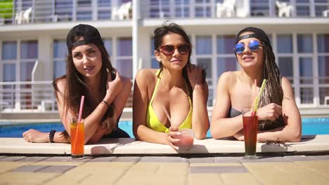 Hermosas-Chicas-Jóvenes-Con-Gafas-De-Sol-Bebiendo-Cócteles-Relajándose-Junto-A-La-Piscina