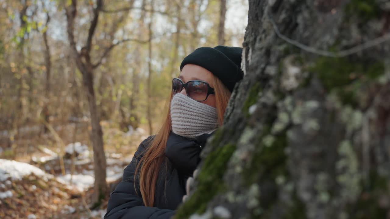 Caucasian redhead woman peeking from behind birch, masked and bundled in scarf and beanie, sunglasses hiding expression, mossy trunk closeup, snowdusted ground, lingering suspense suggesting stealthy