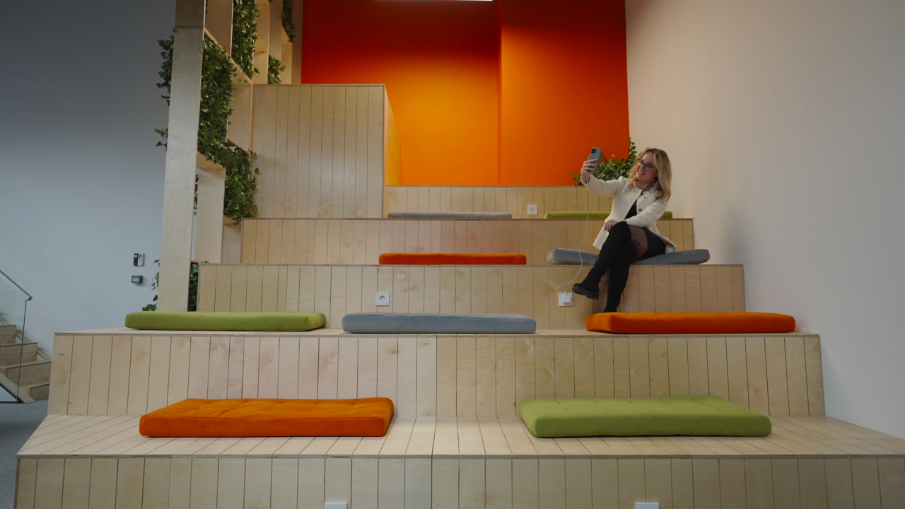 Female Marketing Manager Sitting On Tiered Seating Holding Smartphone For Selfie Orange Accent Wall Wooden. Creative