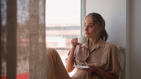 blonde woman enjoys view outside window drinking coffee