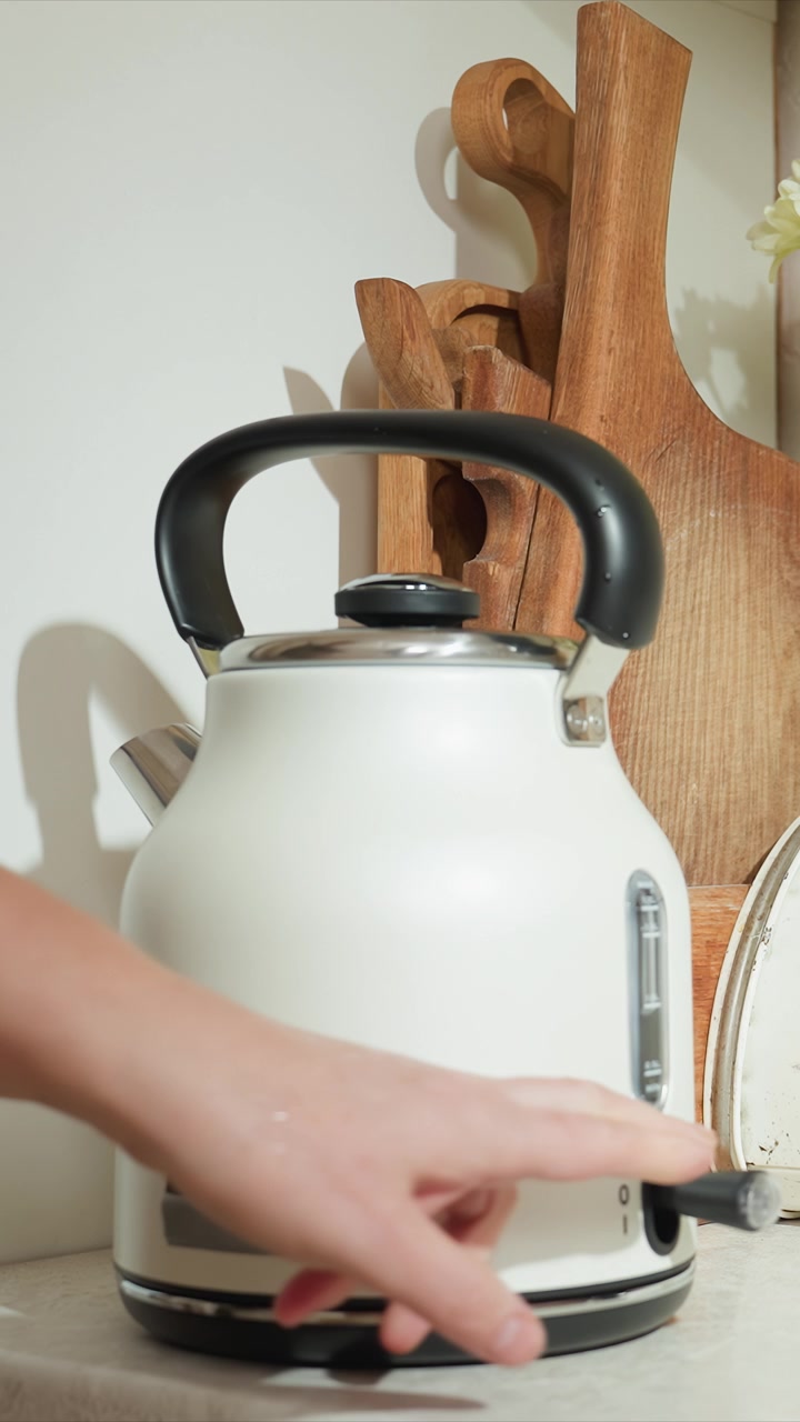 Una mano interagisce con un bollitore elettrico bianco sollevandolo per il suo manico nero. Utensili da cucina in legno sono presenti. La scena è ravvicinata per mostrare i dettagli sia dell'elettrodomestico che del bollitore.