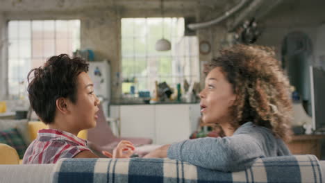 two laughing mixed race girl friends hanging out on couch sharing gossip