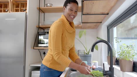 happy african american woman rinsing vegetables in kitchen, slow motion