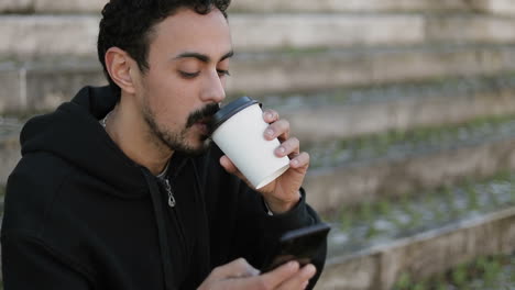 young arabic man with dark curly hair and beard in black hoodie sitting on stairs outside