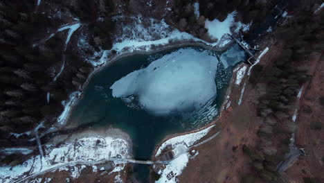 Aerial-view-looking-down-on-a-frozen-mountain-view-surrounded-by-a-winter-forest