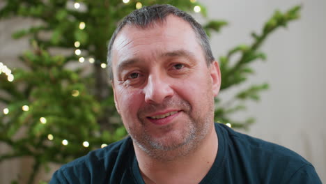 portrait view of cheerful middle aged man smiling warmly with decorated christmas tree lights glowing softly in background creating cozy joyful holiday atmosphere capturing heartfelt