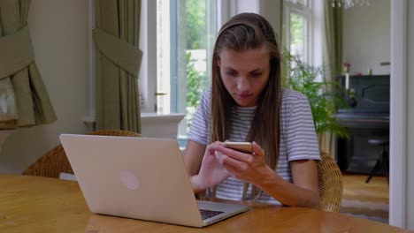 young woman sits at table at home using phone and laptop, slow motion