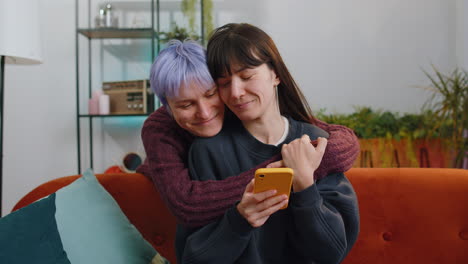 Two-young-lesbian-women-family-married-couple-working-on-smartphone,-shopping-at-home-living-room