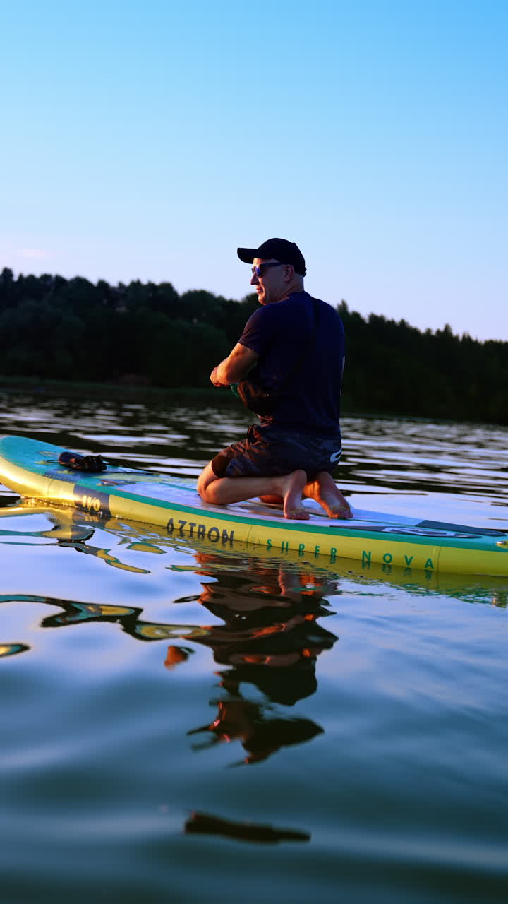 Sportsman kneeling on the inflatable sup board floats by the river. Man uses an oar rowing to the left and to the right. Vertical video.