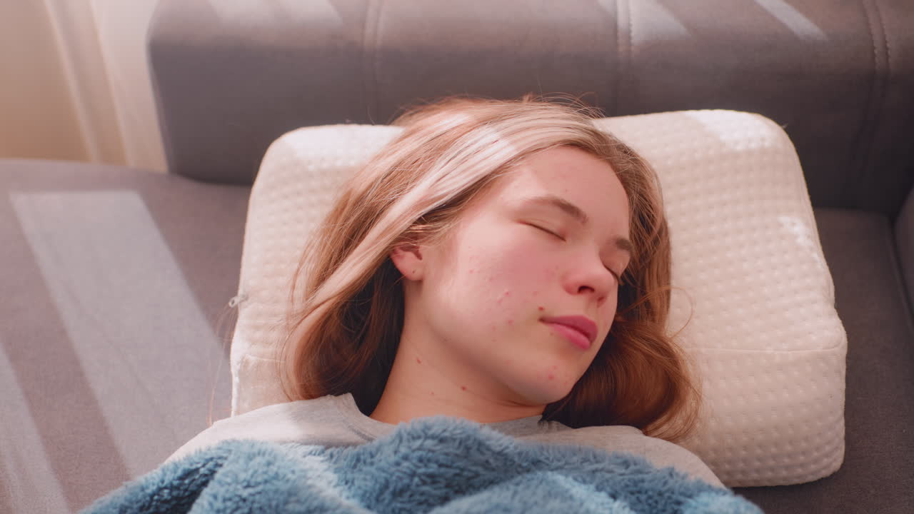 Aerial view of lazy woman sleeping peacefully on couch during day wrapped in fluffy blue blanket, resting head on pillow with sunlight streaming through curtains