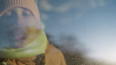 a young girl in a pink knit hat and scarf blowing air onto a foggy car window, creating condensation. her face is pressed against the glass