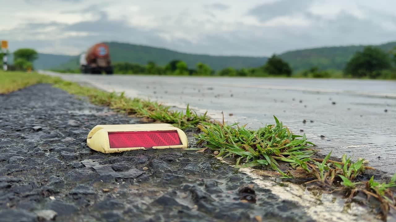 Static close-up of a wet road reflector marker on asphalt after rain, with a blurred truck driving away in the background on a countryside highway