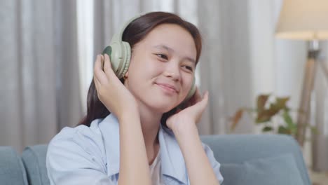 close up of asian teen girl with headphones listening to music and dancing while sitting on sofa in the living room at home