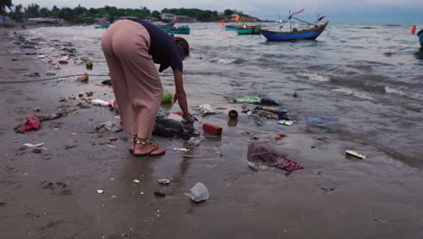 a woman picking up environmental waste from beach full of plastic and debris along coast