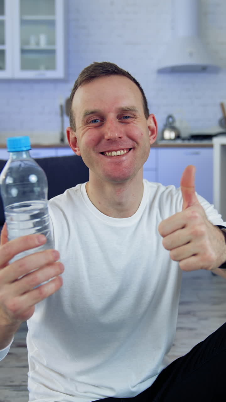 Smiling young man with a bottle of water at home. Young man proposing fresh water after the workout and looking on camera indoors. Vertical video