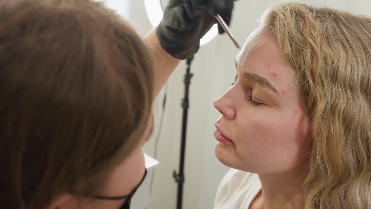 Beautician wearing black gloves applies and blends cream on both eyebrows of client with closed eyes in beauty salon, using pink-handled brush. Ring light visible in softly blurred background